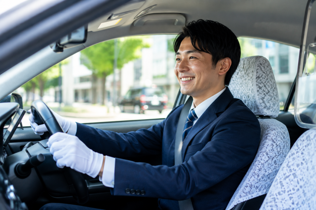 タクシー車内で笑顔で運転するタクシードライバーの仕事風景。地域のお客様を安全・快適に目的地まで送迎する様子をイメージした求人用画像。