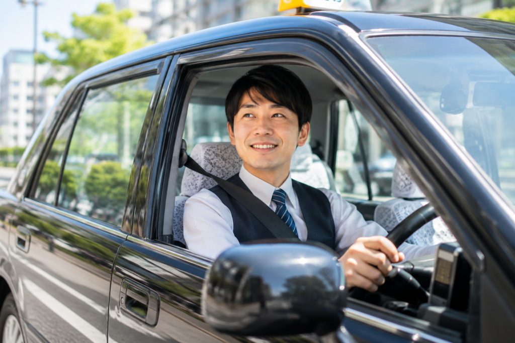 川崎市内を走るタクシーの運転席で笑顔で乗務するタクシードライバーの仕事風景イメージ